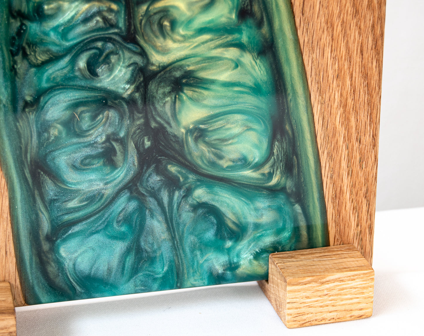 Primer plano en ángulo de una tabla de servir de roble con resina epoxy verde, mostrando la textura ondulada de la resina y un soporte de madera.