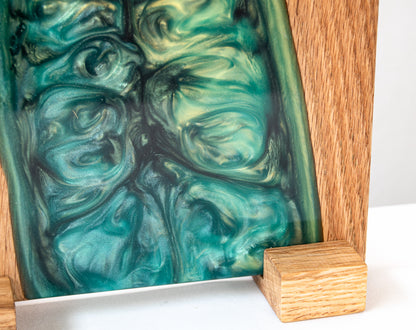 Primer plano en ángulo de una tabla de servir de roble con resina epoxy verde, mostrando la textura ondulada de la resina y un soporte de madera.