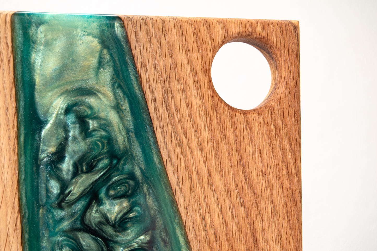 Primer plano de una tabla de servir de roble con banda diagonal de resina epoxy verde y orificio para asa, sobre fondo claro.