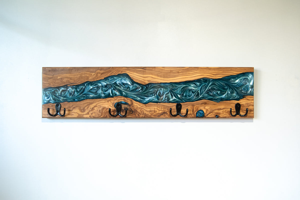 Perchero de pared en madera de olivo con veta de resina epoxi azul turquesa metalizado y cuatro ganchos negros, fotografiado sobre fondo blanco