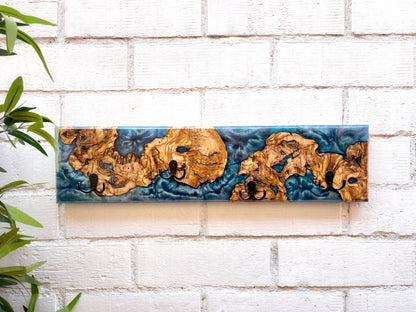 Perchero de pared artesanal en madera de olivo con resina epoxi en tonos azul teal y cuatro ganchos negros, sobre pared de ladrillo blanco