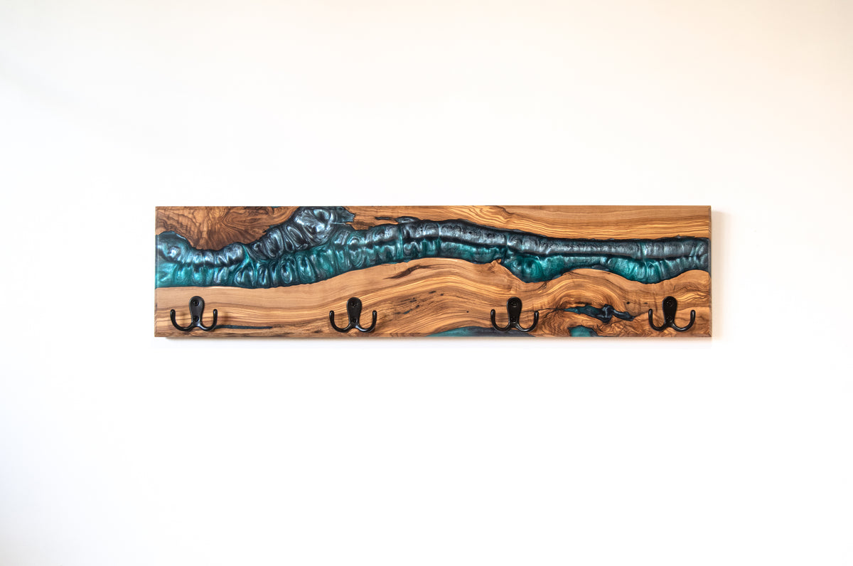 Perchero de pared artesanal en madera de olivo con acabado en resina epoxi azul turquesa metalizado y cuatro ganchos negros, sobre pared blanca lisa