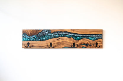 Perchero de pared artesanal en madera de olivo con acabado en resina epoxi azul turquesa metalizado y cuatro ganchos negros, sobre pared blanca lisa