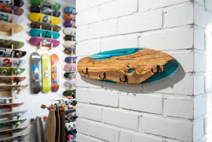 Perchero de pared en forma de tabla de skate, madera de olivo con resina epoxi azul turquesa y cuatro ganchos negros, junto a pared con tablas de skate