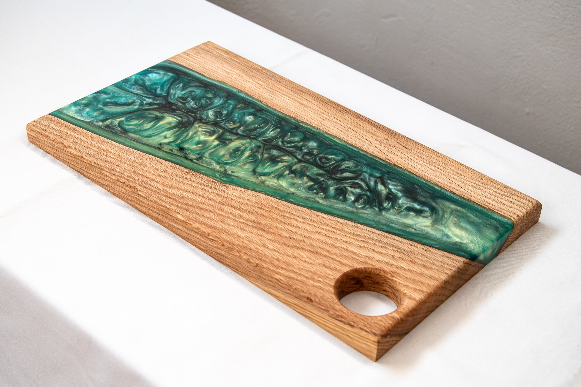Tabla de servir de madera de roble con banda diagonal de resina epoxy verde y orificio para asa, fotografiada sobre una mesa blanca