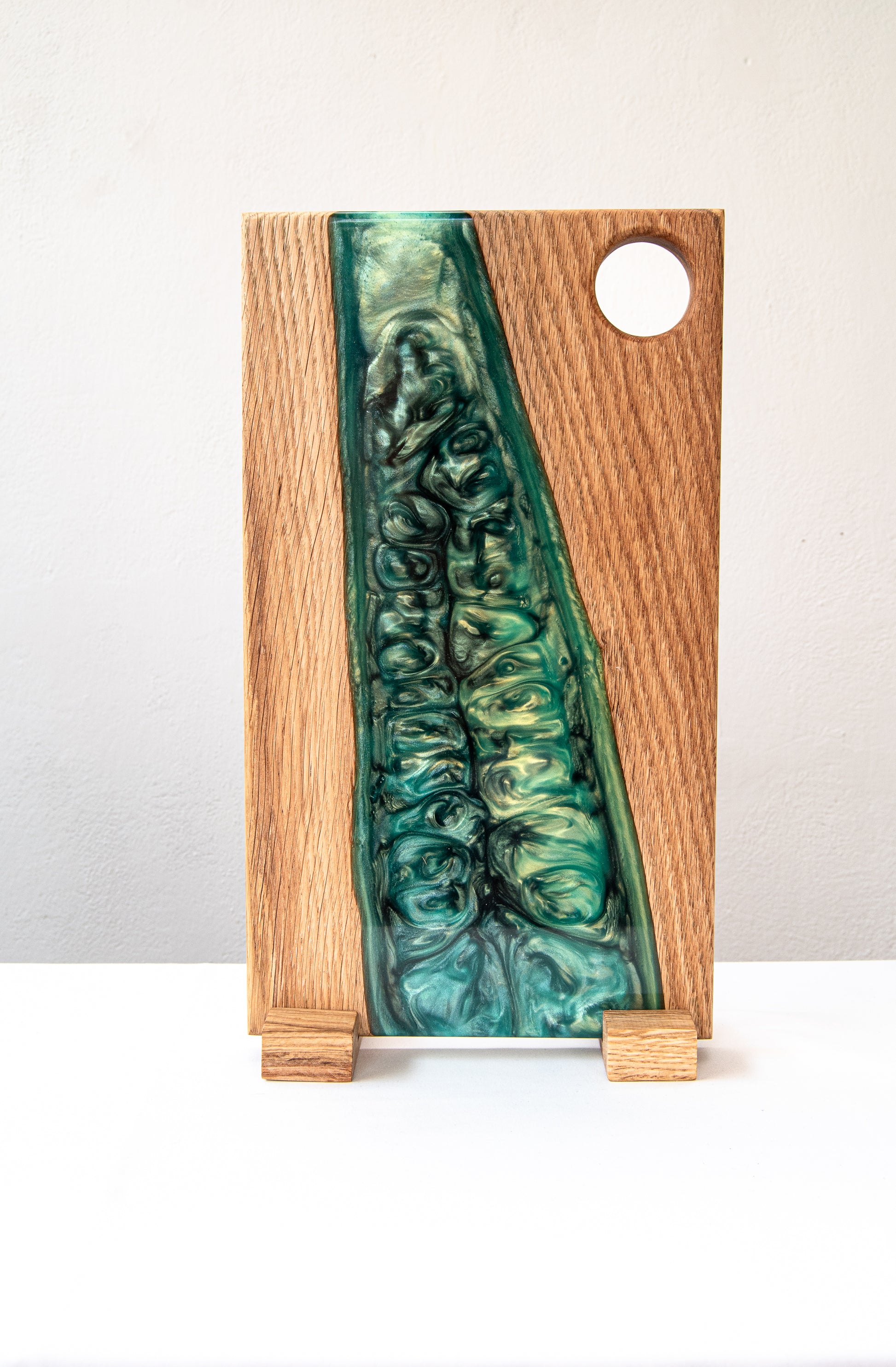 Vista vertical de una tabla de servir de madera de roble con resina epoxy verde y orificio para asa, de pie sobre fondo claro